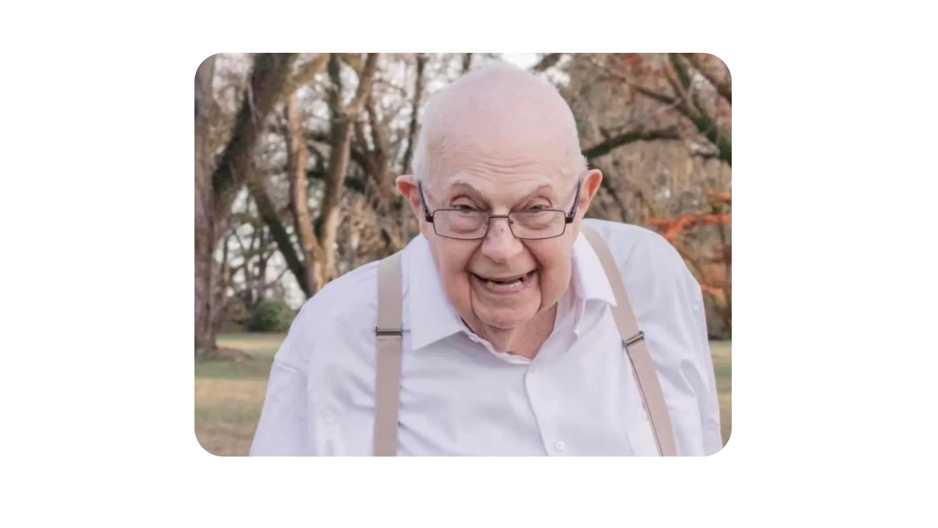 Elderly man with glasses and smiling outdoors, wearing a white shirt, with bare winter trees in the background.