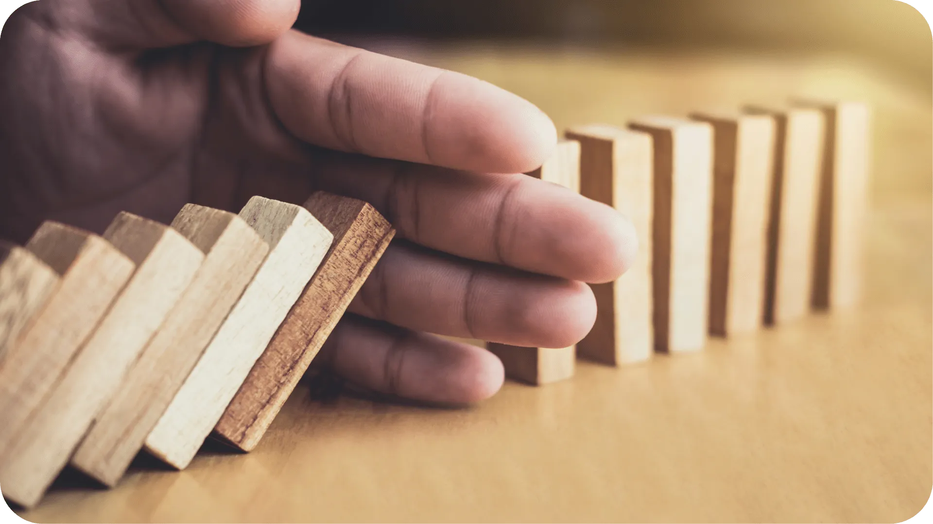 Wooden blocks stood up in a line like dominoes. There is a hand in the way, stopping the falling ones from knocking the rest over.